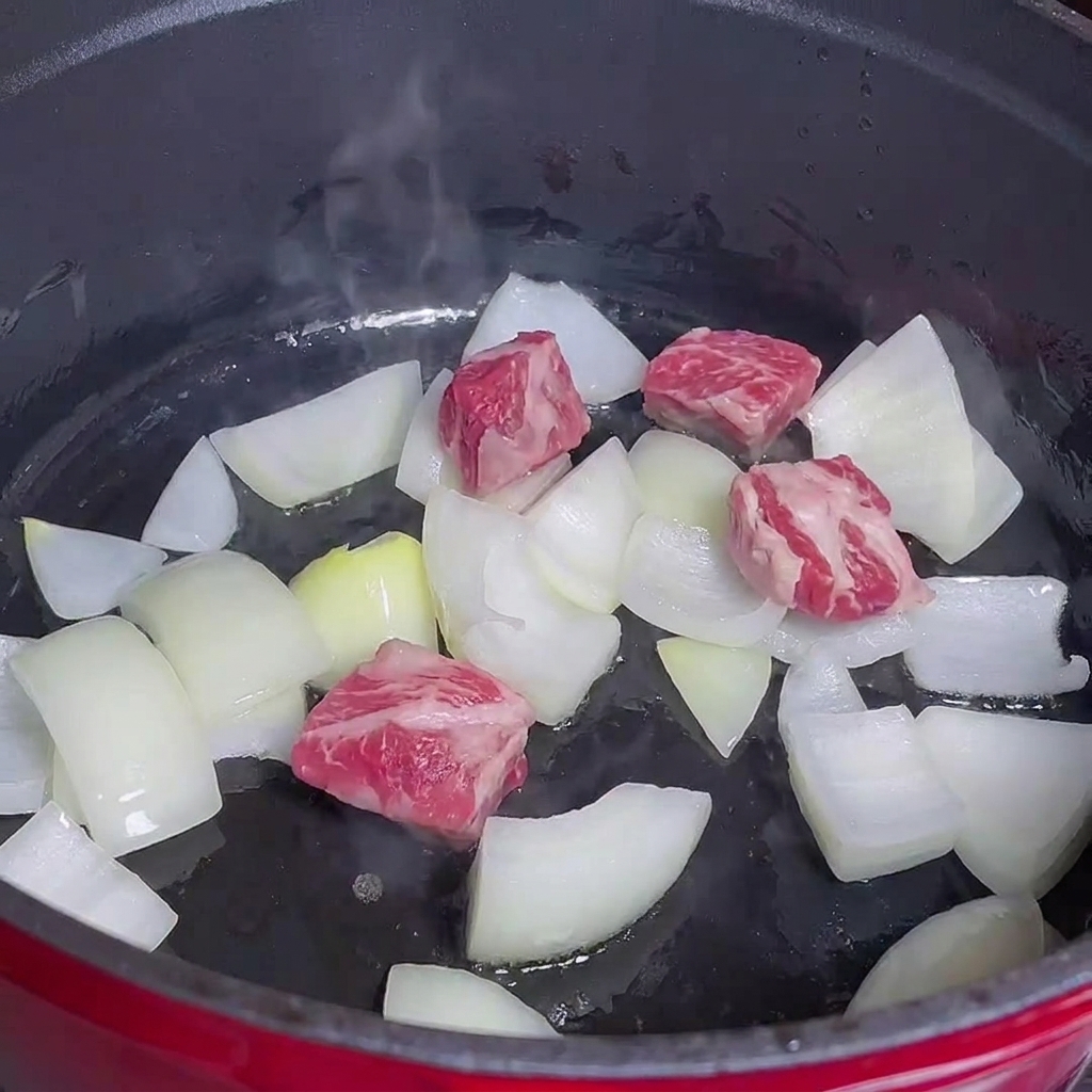 Fleischwürfel garen neben Zwiebeln in einem roten Topf.