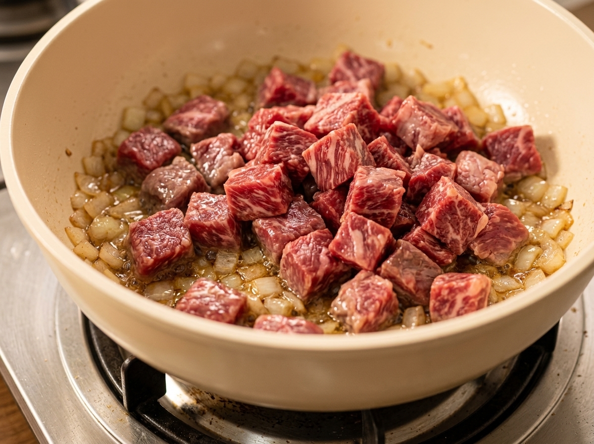 Gewürfeltes Rindfleisch wird in eine Pfanne mit angeschwitzten Zwiebeln und gewürfeltem Gemüse gegeben.