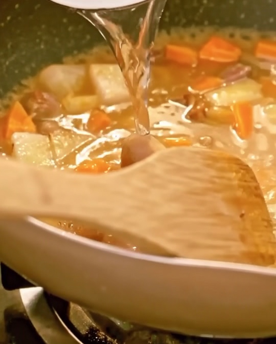 Wasser wird in eine Pfanne mit Rindfleisch, Gemüse und schmelzendem Curry gegossen, ein Holzspatel ist sichtbar.