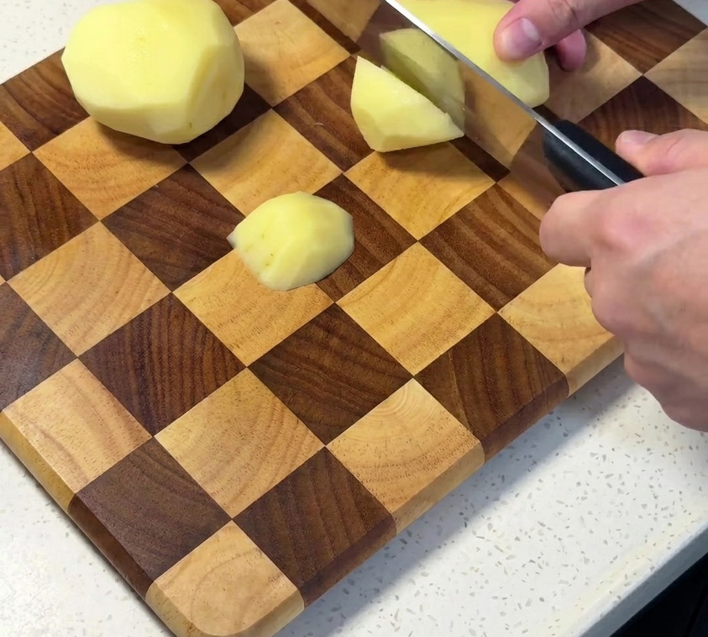 Geschälte rohe Kartoffeln werden auf einem Holzschneidebrett in Würfel geschnitten.