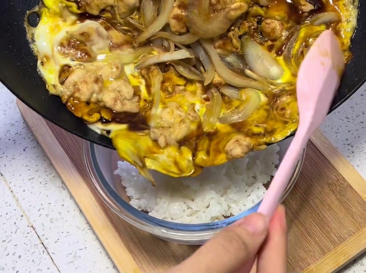 Nahaufnahme der Hähnchen-, Ei- und Zwiebelmischung, die aus der Pfanne auf eine Schale mit dampfendem weißen Reis umgefüllt wird.