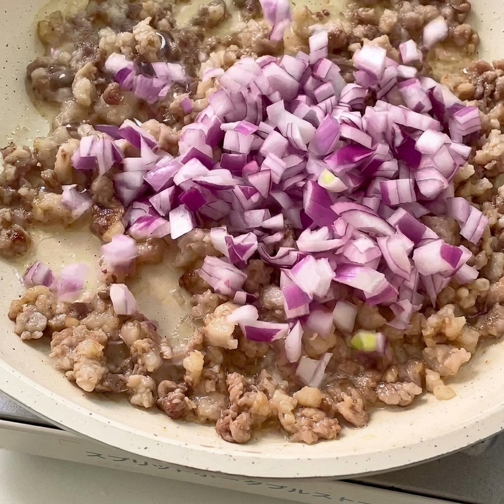 Gehackte rote Zwiebel und Knoblauch werden mit dem gebräunten Fleisch in der Pfanne angebraten.