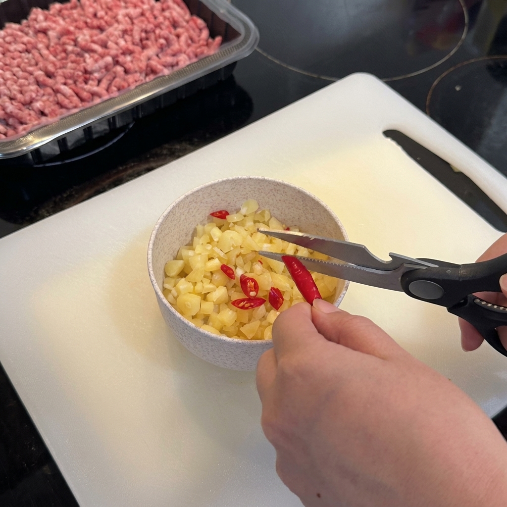 Küchenschere schneidet leuchtend rote Thai-Chilis direkt in eine Schüssel mit gehacktem Knoblauch.