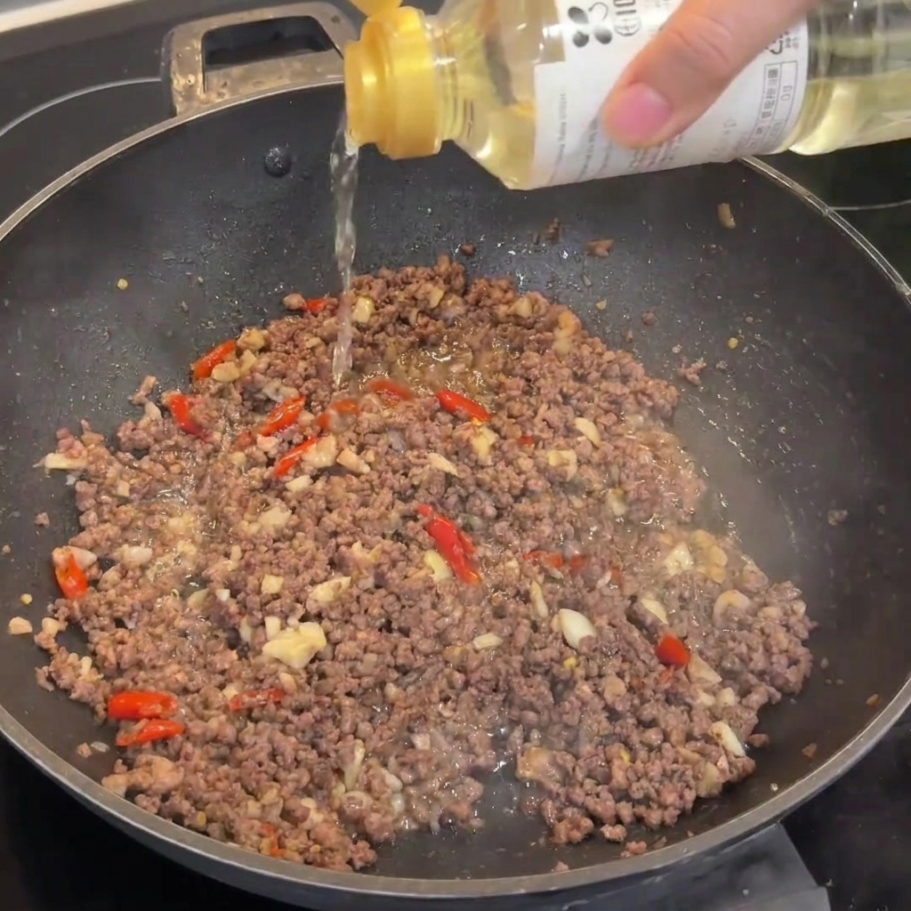 Ein Spritzer Reiswein wird in einen Wok mit gebräuntem Rinderhackfleisch gegossen.