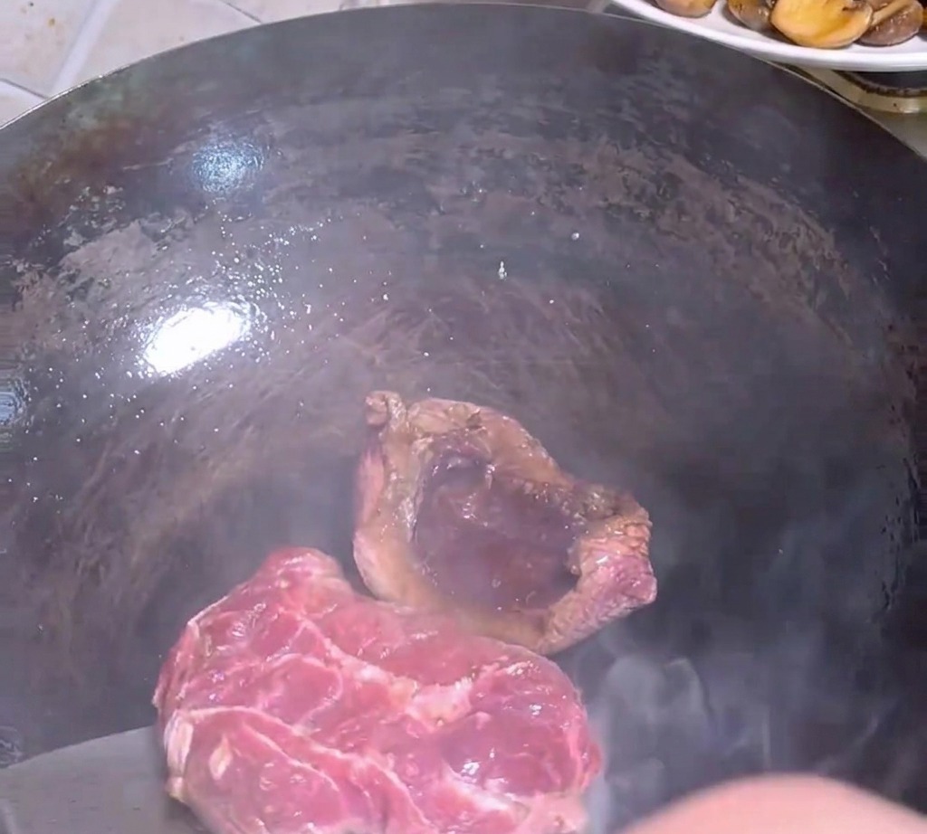 Ein Metallspatel wendet ein teilweise angebratenes Steak in einem heißen, dunklen Wok.
