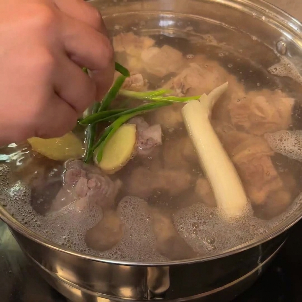 Eine Nahaufnahme von großen weißen Zwiebelstücken und Frühlingszwiebeln, die zu den blanchierten Schweineknochen in den Topf gegeben werden.