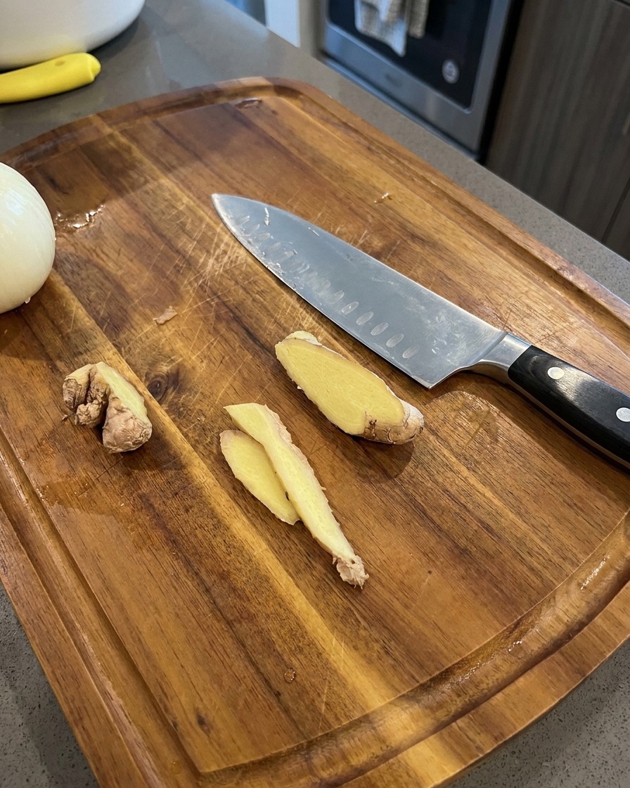 Dünne Ingwerscheiben liegen auf einem Holzschneidebrett neben einem Kochmesser und einer Zwiebelhälfte.