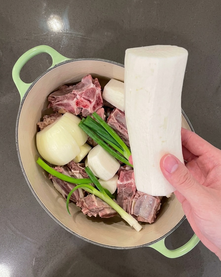 Eine Hand legt große Stücke weißen Rettich in den Topf neben das Fleisch und die Aromaten.