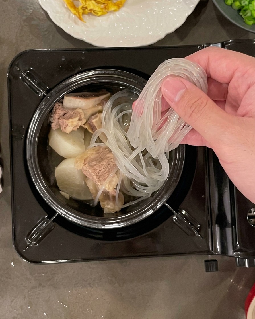 Eine Hand legt ein Bündel eingeweichte, durchscheinende Glasnudeln in einen mit Rindfleisch und Rettich gefüllten Steintopf.