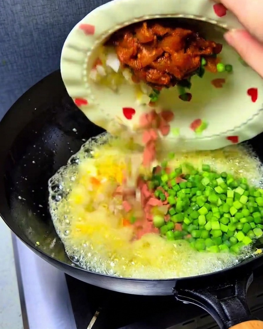 Gehacktes Kimchi, gewürfelter Schinken, Hähnchen und grüne Knoblauchsprossen werden von einem Teller in den Wok mit den Eiern gegossen.