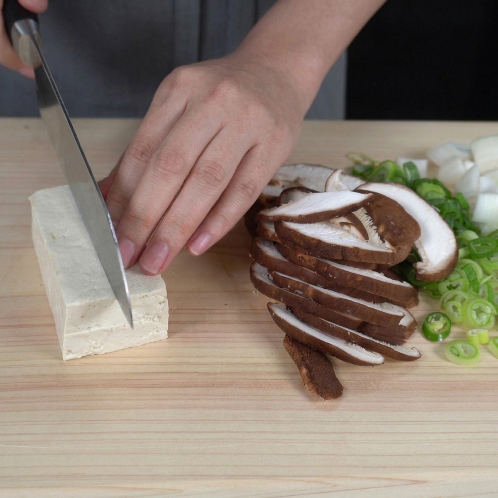 Hände schneiden einen Block festen weißen Tofu auf einem Schneidebrett in gleichmäßige, mundgerechte Würfel.