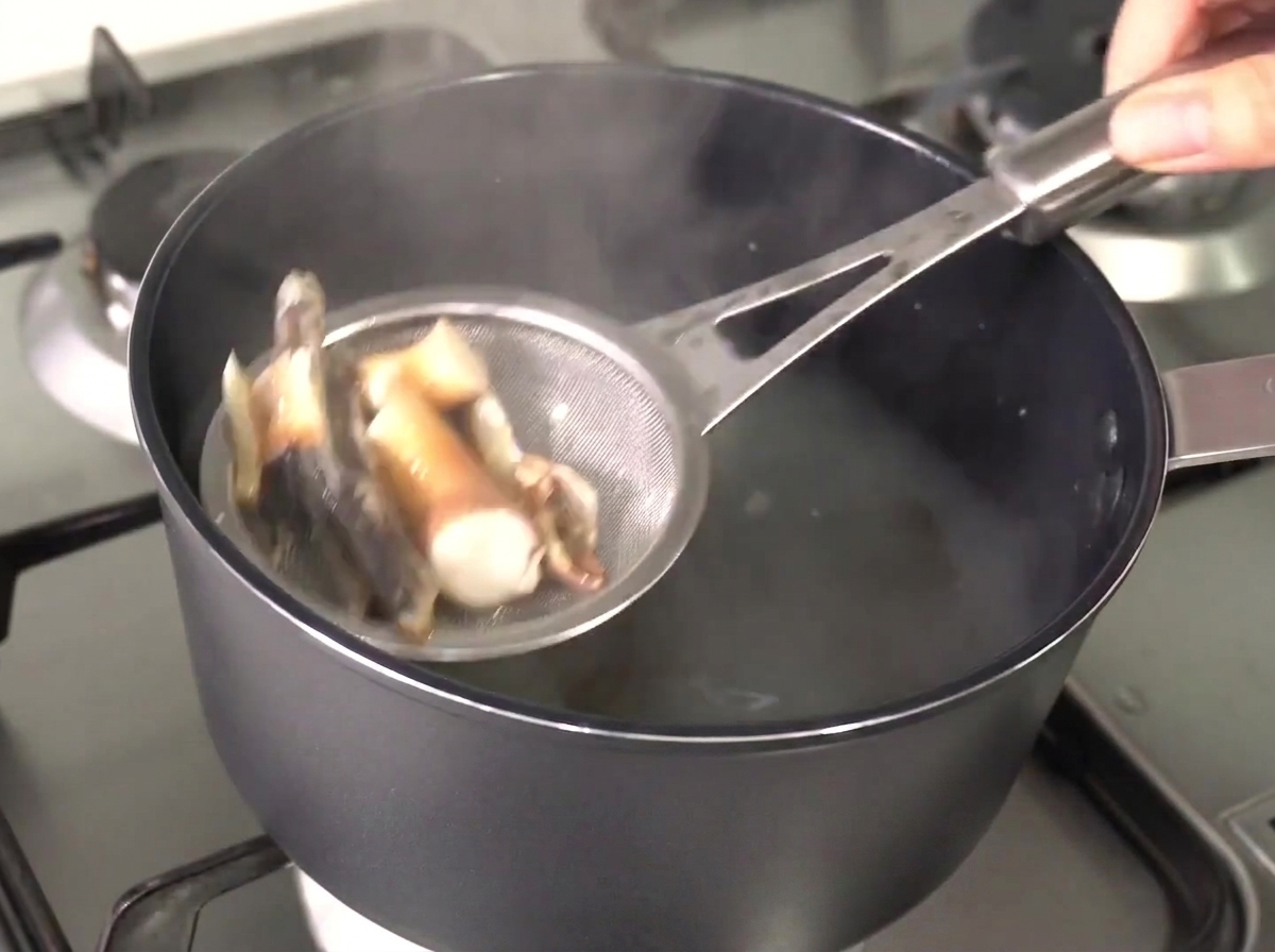 Ein Metallsieb hebt weich gekochte Sardellen und Seetang aus der heißen, klaren Brühe.