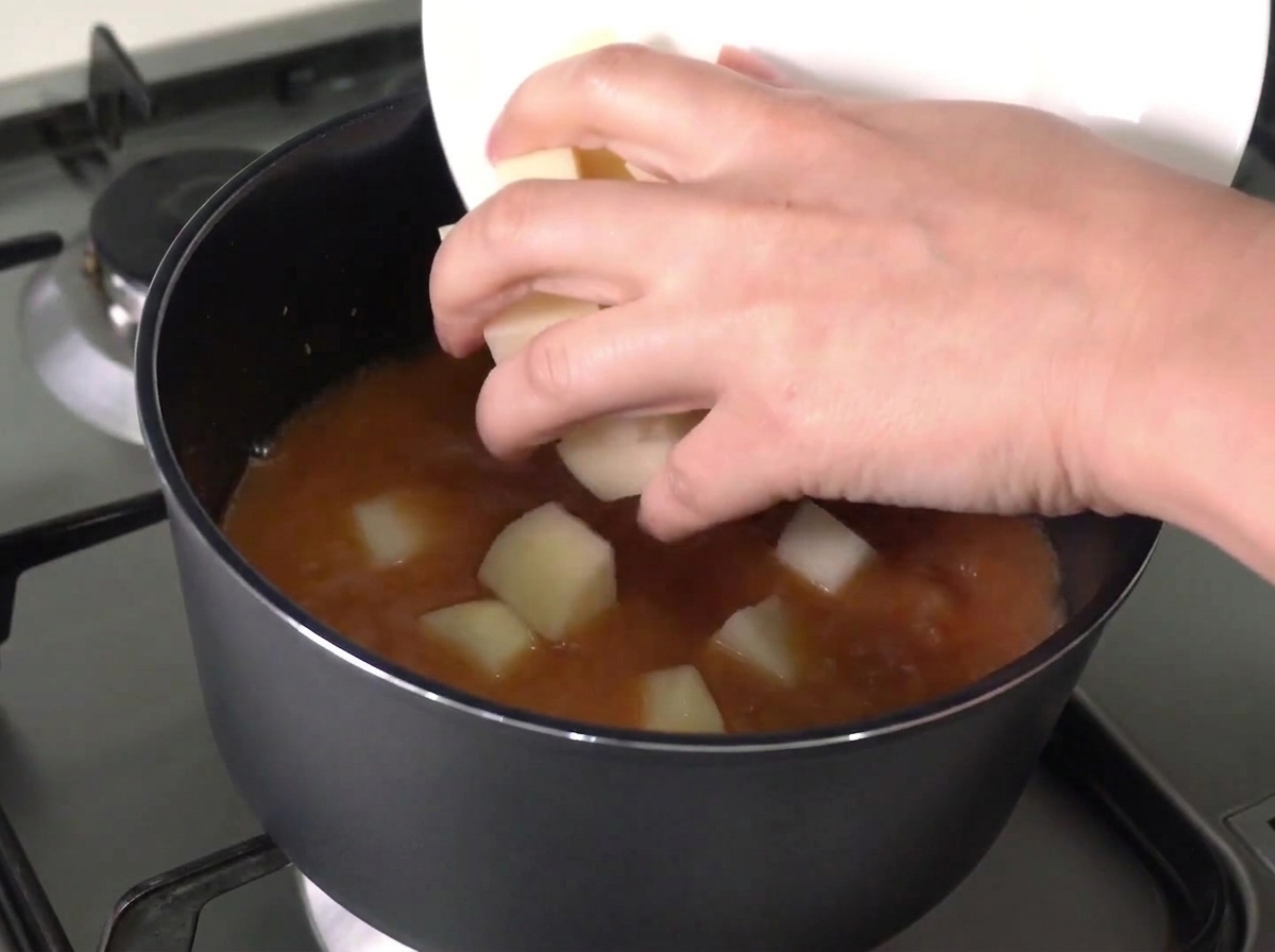 Gehackte Kartoffeln und Zwiebeln werden von einem weißen Teller in einen Topf mit kochender roter Brühe geschoben.