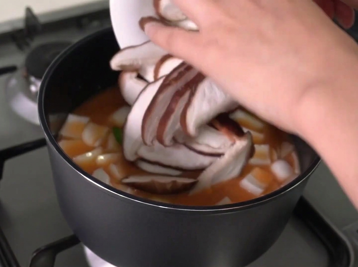 In Scheiben geschnittene Shiitake-Pilze werden von einem weißen Teller in einen Topf mit kochendem Eintopf gegeben.