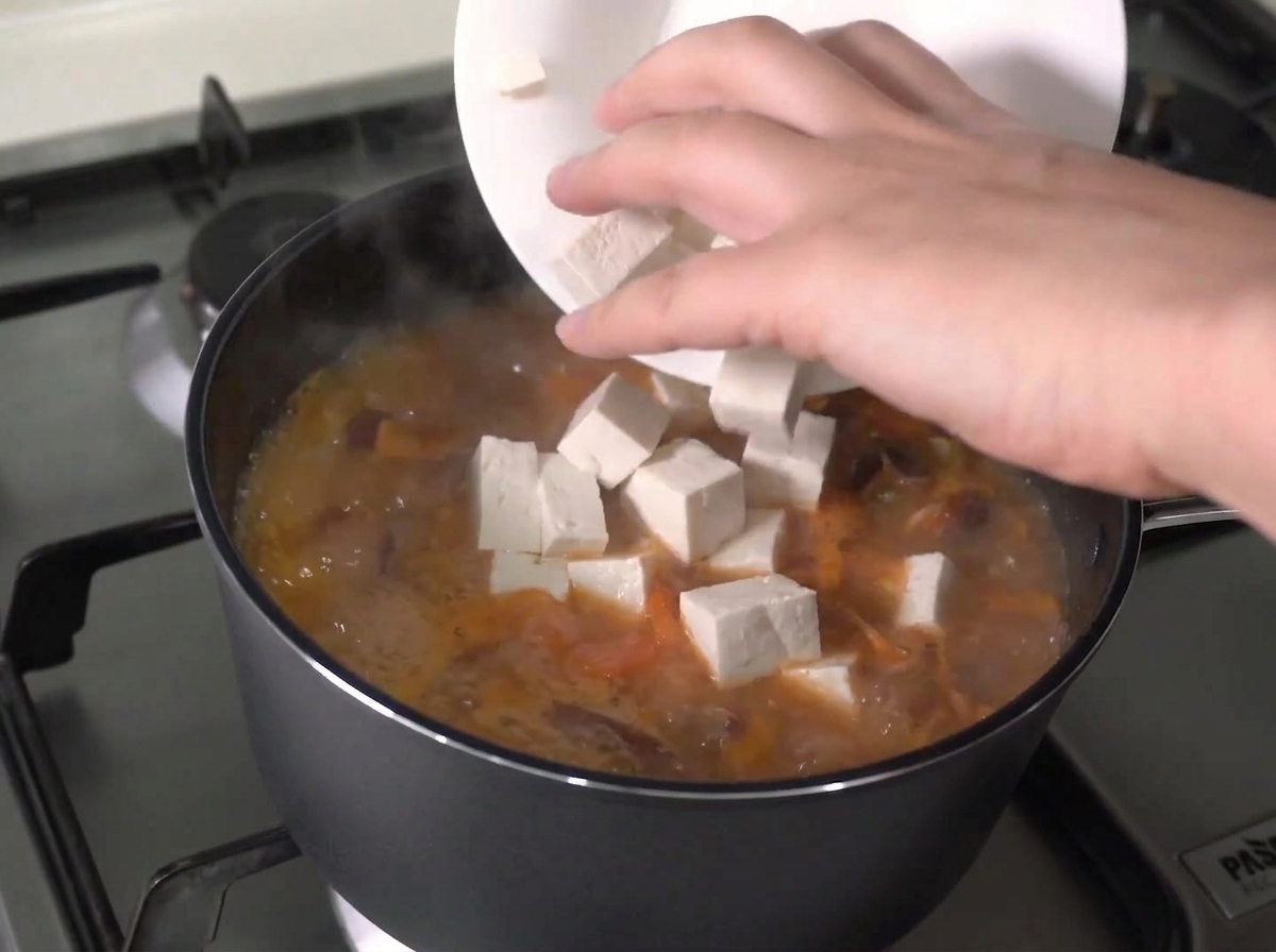 Frische Tofuwürfel werden von einem weißen Teller in einen stark kochenden Eintopf gegeben.