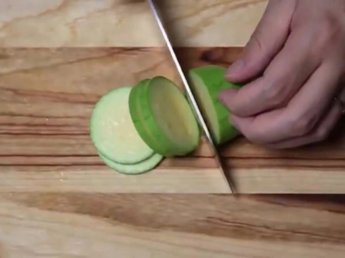 Eine grüne Zucchini wird auf einem Holzschneidebrett in gleichmäßige Halbmonde geschnitten.