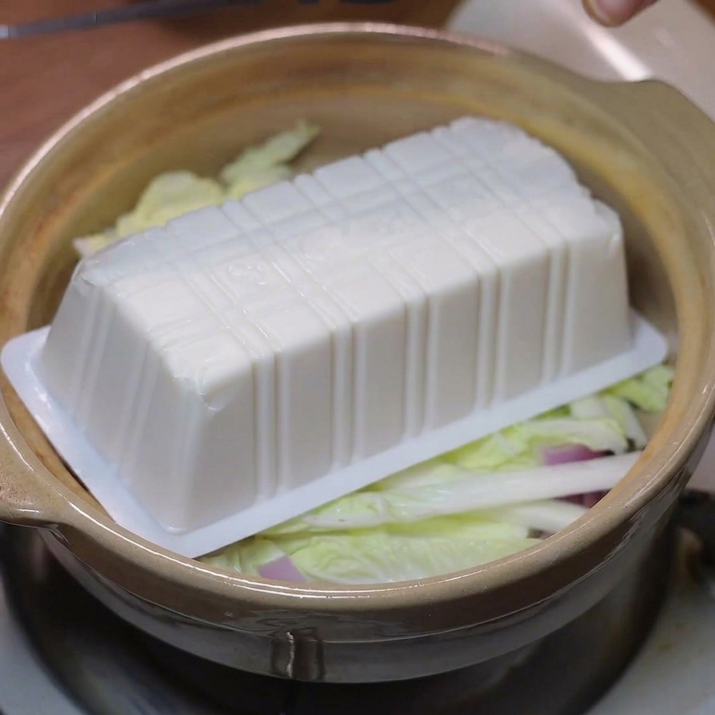 Stücke von weichem Tofu werden auf das Gemüse im Tontopf gelegt.
