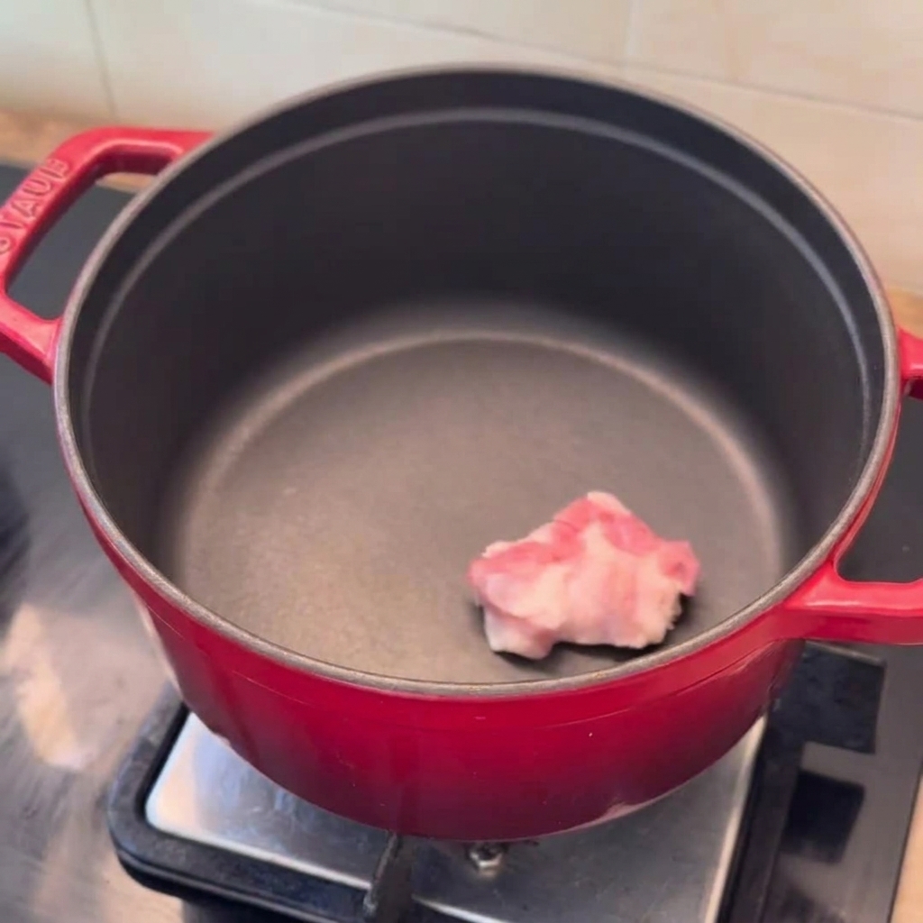 Schweinebauchscheiben, Zwiebelspalten und gehackter Knoblauch liegen bereit in einem vorgeheizten roten Topf.