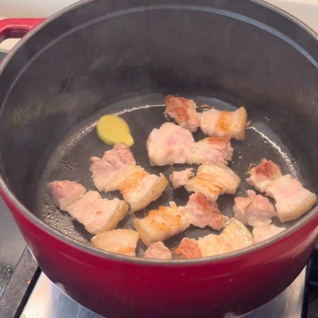 Goldbraune Schweinebauchstücke werden in einem roten Topf angebraten.