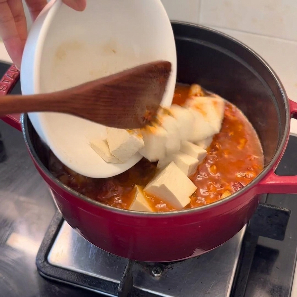 Frische Tofuwürfel werden vorsichtig aus einer weißen Schale in einen tiefroten Topf mit blubberndem Kimchi-Schweinefleisch-Eintopf gegeben.