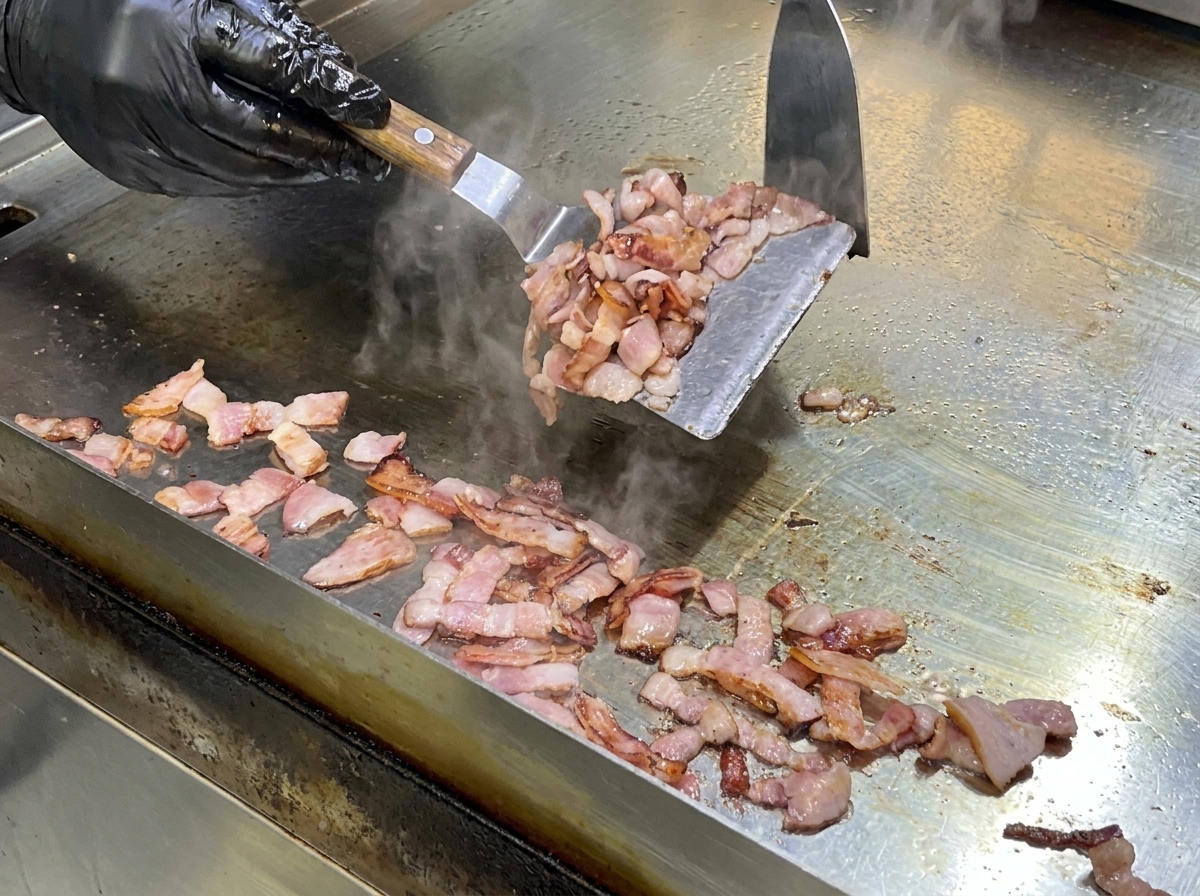 Ein Metallspatel rührt Baconstücke auf einer heißen Grillplatte, um das Fett auszulassen.
