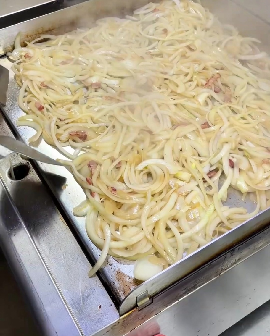 Ein Spatel rührt einen großen Haufen weich werdender weißer Zwiebeln gemischt mit Baconstücken auf einer dampfenden Metallplatte.