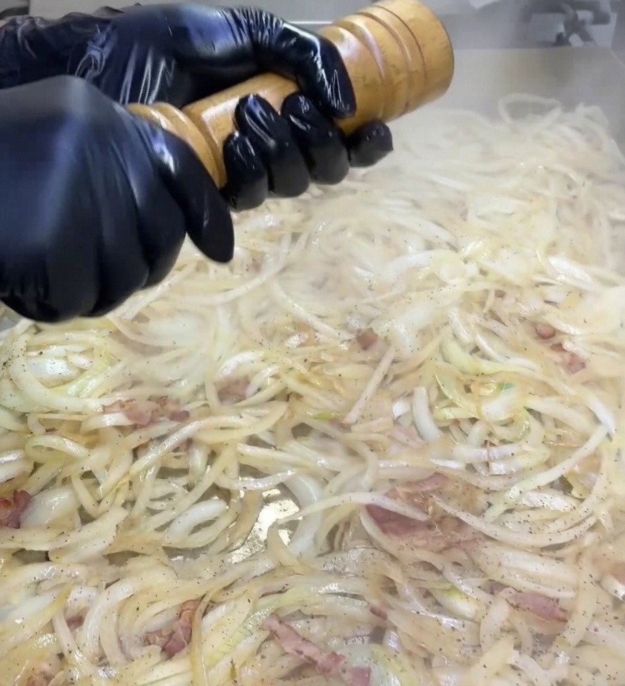 Hände in schwarzen Handschuhen drehen eine hölzerne Pfeffermühle über einer mit Zwiebeln und Bacon bedeckten Grillplatte.