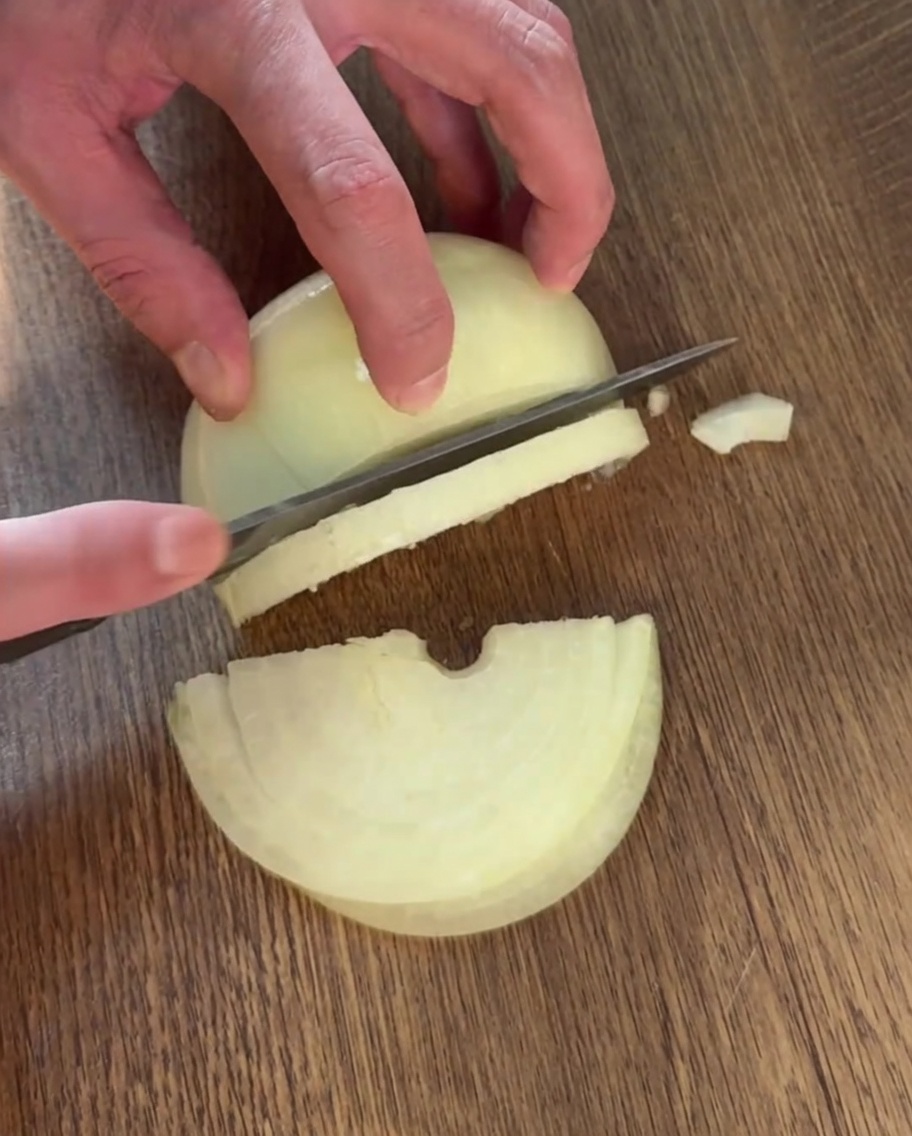 Eine Hand schneidet mit einem Messer eine Zwiebel in dünne Streifen auf einem Holzschneidebrett.