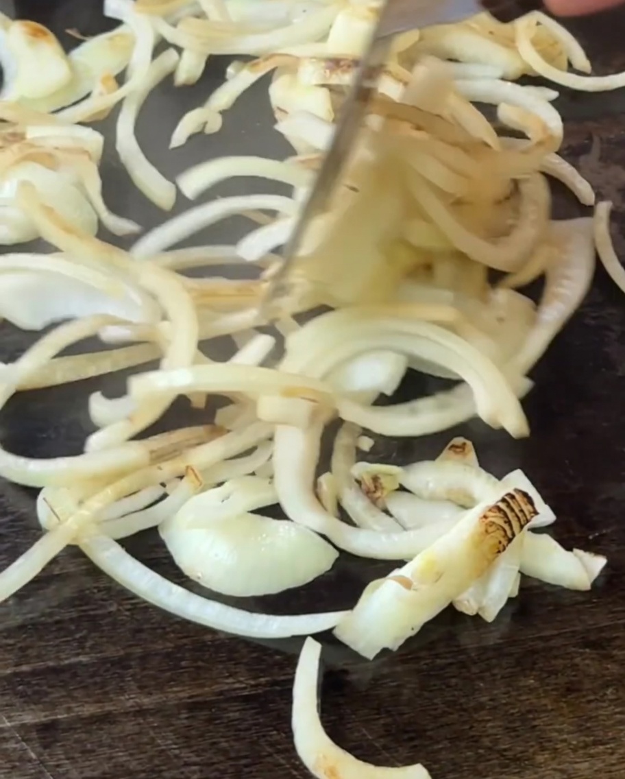 Zwiebeln werden auf der Grillplatte mit einem Pfannenwender gewendet, während sie weich werden.