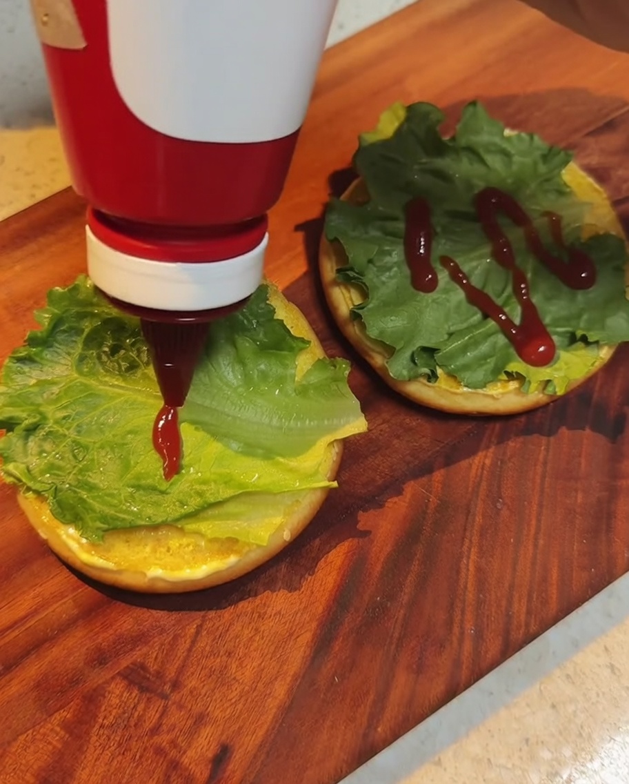 Roter Ketchup wird aus einer Flasche über die Salatblätter auf dem Brötchen geträufelt.