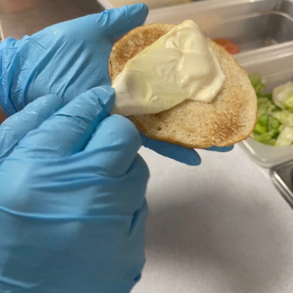Ein Kunststoffschaber verteilt Mayonnaise auf einem getoasteten Burgerbrötchen.