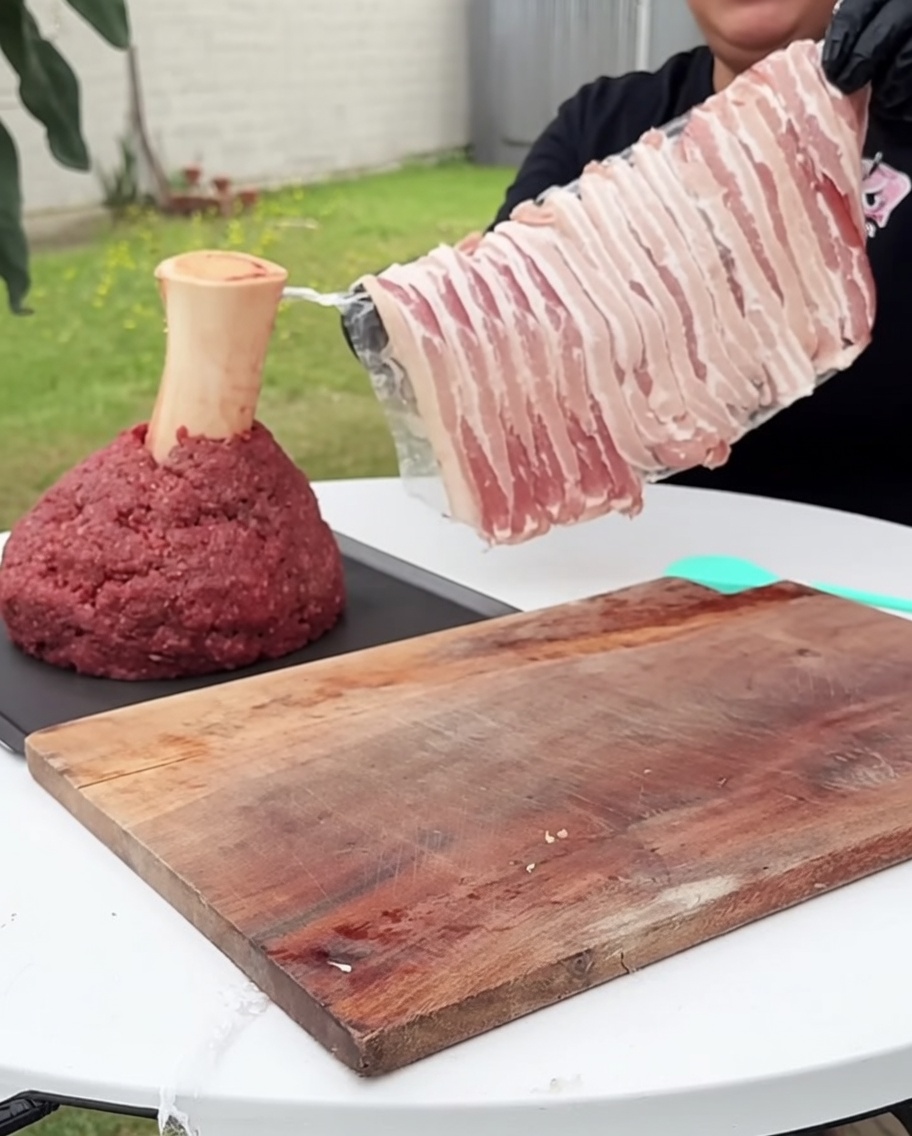 Ein quadratischer Block Rinderhackfleisch, der vorsichtig von allen Seiten mit rohen Speckstreifen umwickelt wird.