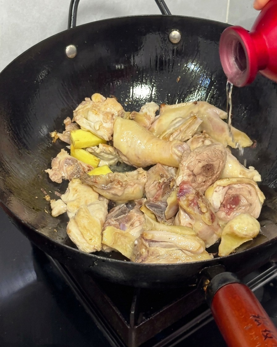 Reiswein wird aus einer roten Flasche in einen Wok mit gebräunten Hühnerstücken und Ingwer gegossen.