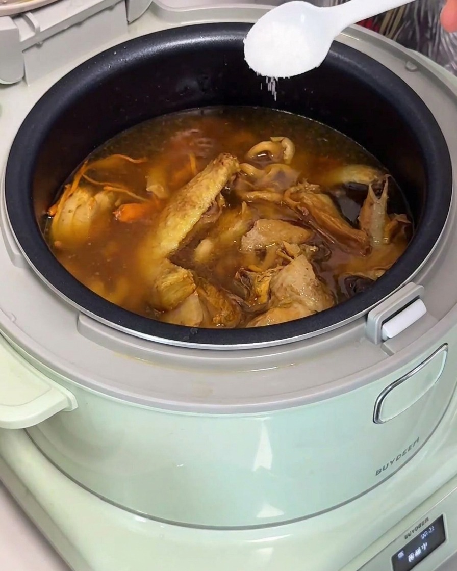 Ein weißer Löffel gibt Salz in einen fertigen Topf mit goldener Hühner-Pilz-Suppe in einem Reiskocher.