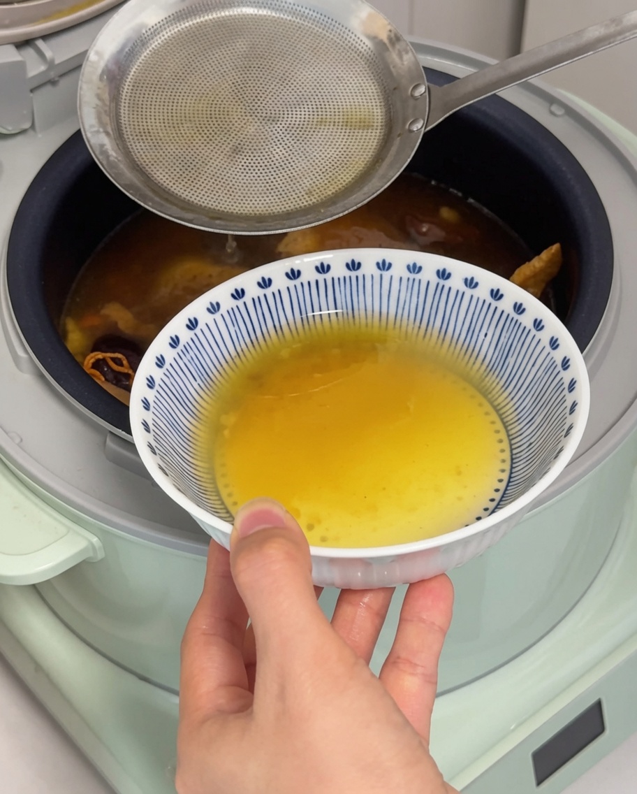 Nahaufnahme einer Metallkelle, die eine Schicht goldenes Hühnerfett von der Oberfläche der Suppe in einem Reiskocher abschöpft.