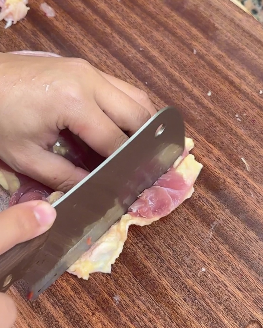 Eine Hand schneidet entbeintes rohes Hähnchenfleisch mit einem Hackbeil auf einem Holzbrett in dünne Streifen.