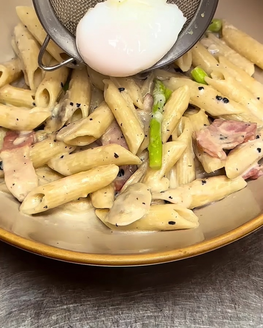Cremige Penne-Pasta wird in einer Schüssel angerichtet und vorsichtig mit einem pochierten Ei belegt.