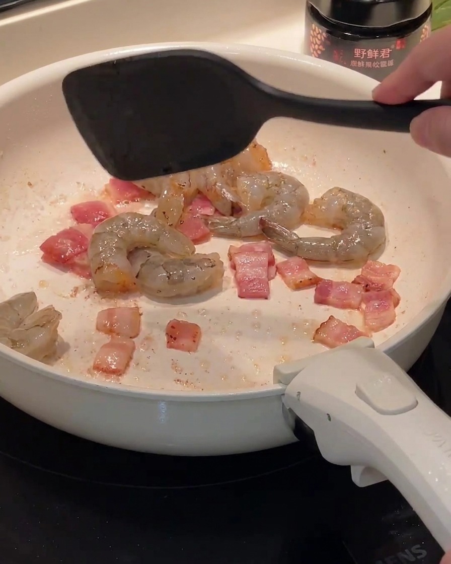 Rohe Garnelen werden in eine Pfanne mit gebratenen Speckstücken gegeben.