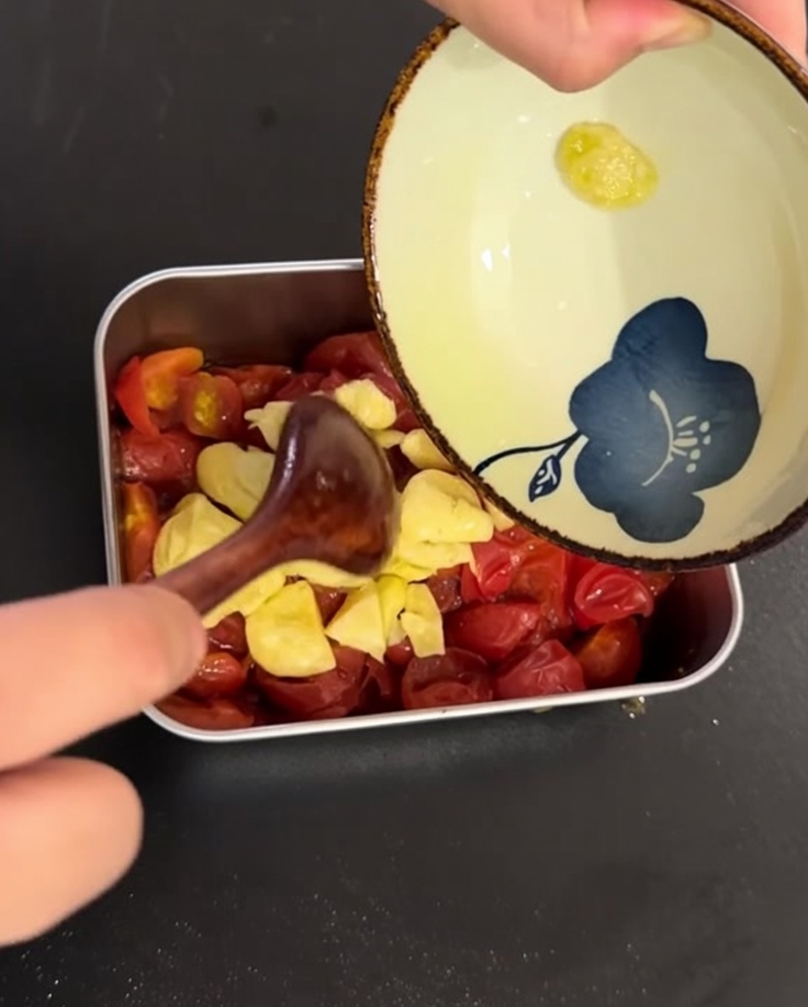 Das mit Knoblauch aromatisierte Olivenöl und der geschnittene Knoblauch werden über die gegarten Kirschtomaten in einer Metallschale gegossen.