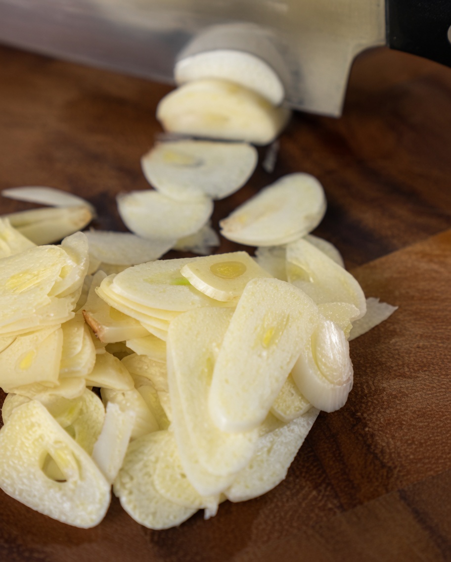 Dünn geschnittene Knoblauchzehen auf einem Holzschneidebrett neben der Klinge eines Kochmessers.