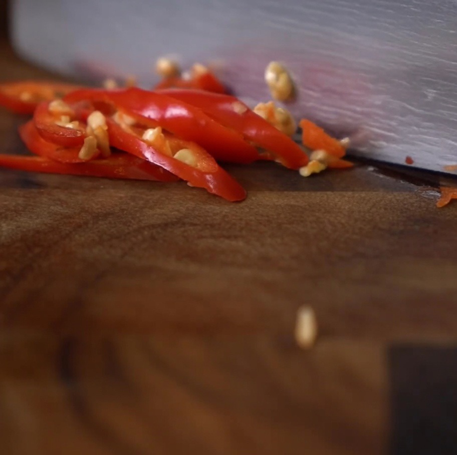 Eine Nahaufnahme eines Messers, das auf einem dunklen Holzschneidebrett leuchtend rote Chilischoten in kleine Ringe schneidet.