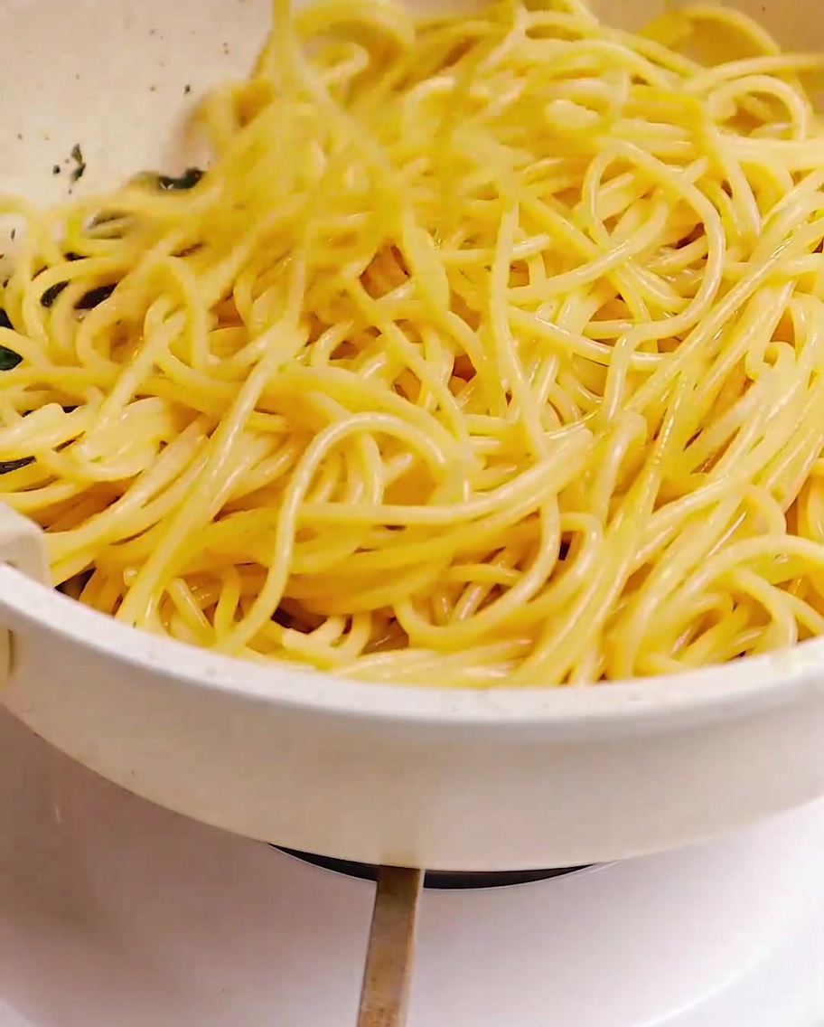 Gekochte Spaghetti werden in einer Pfanne mit Stäbchen in der Speck-, Knoblauch- und Kräutermischung geschwenkt.