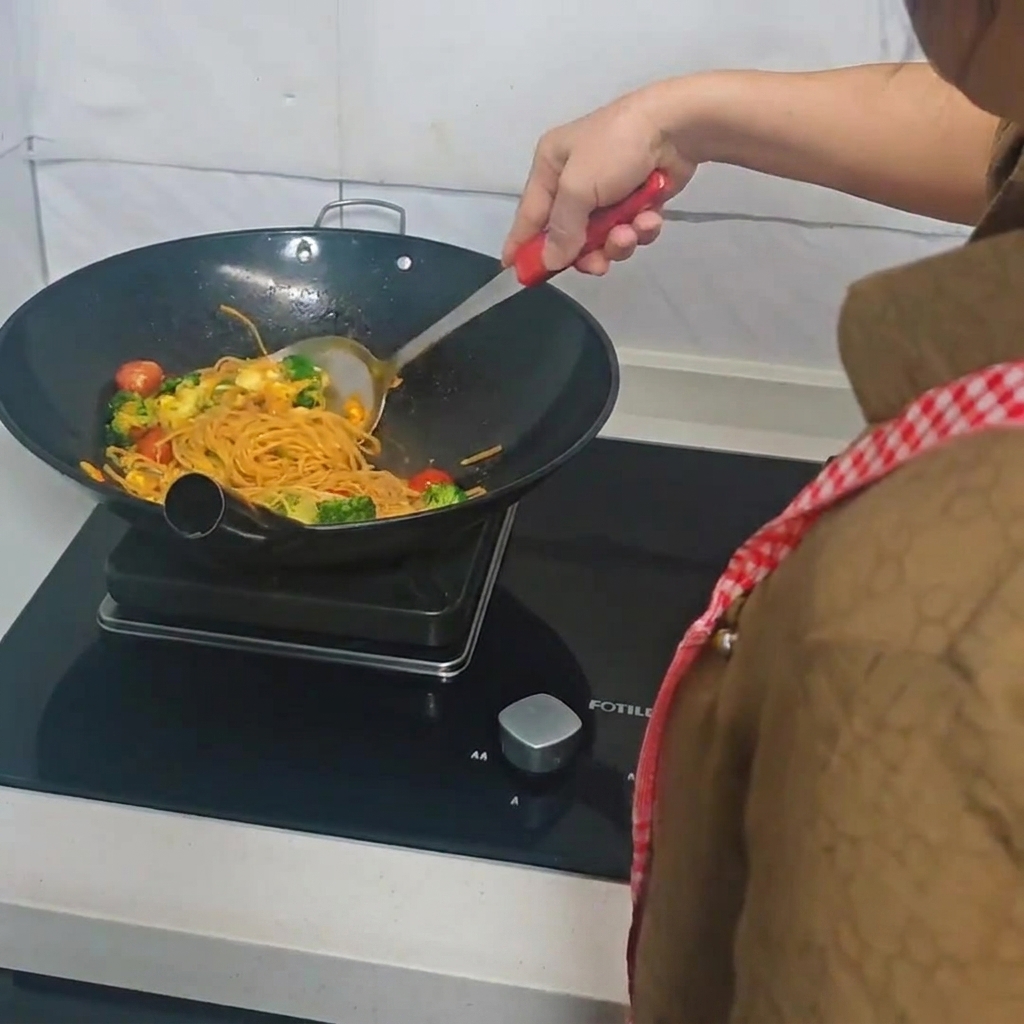Eine Person wendet Spaghetti, Brokkoli, Kirschtomaten und Eier mit einem Spatel im Wok.