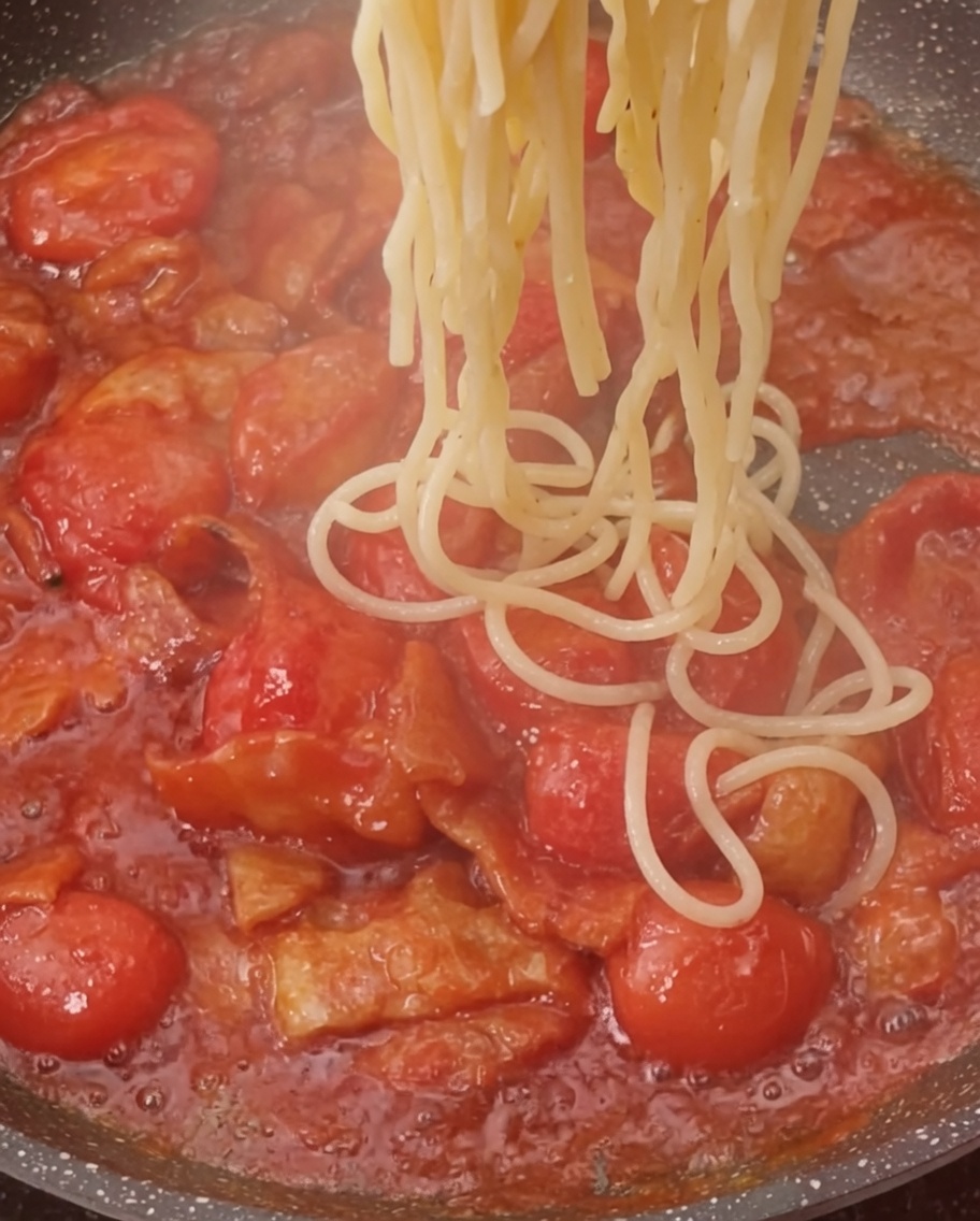 Gekochte Nudeln werden in die Pfanne zur Tomaten-Speck-Sauce gegeben.