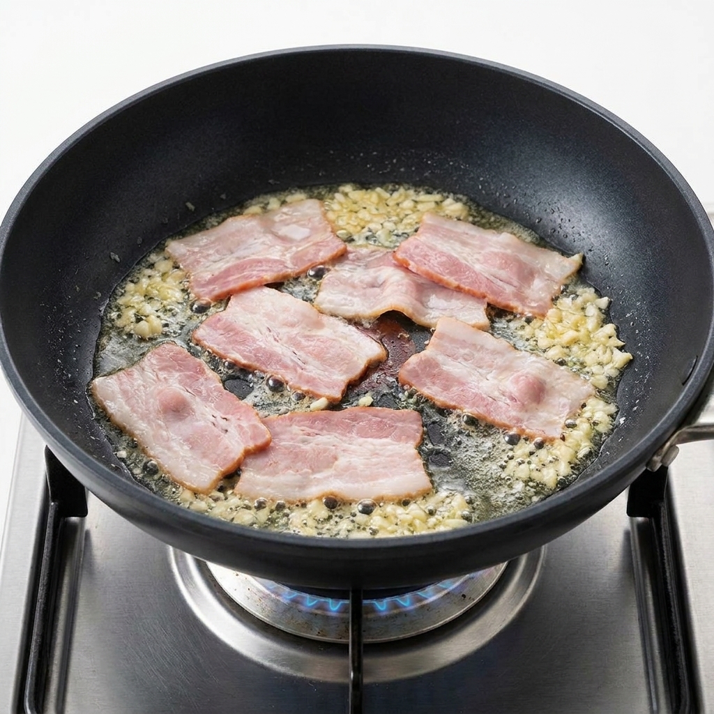 Speckstücke braten in der Pfanne neben duftendem, goldenem Knoblauch.