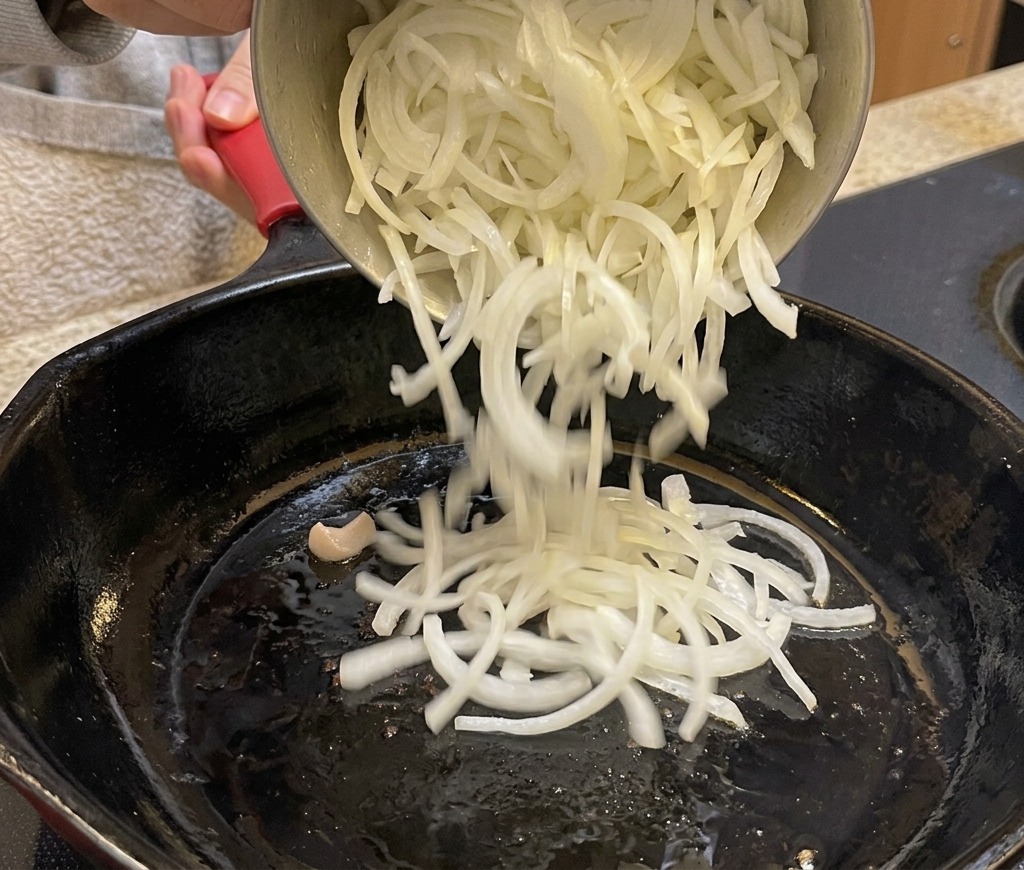 Dünn geschnittene Zwiebeln werden aus einem Topf in eine schwarze Pfanne gegossen.