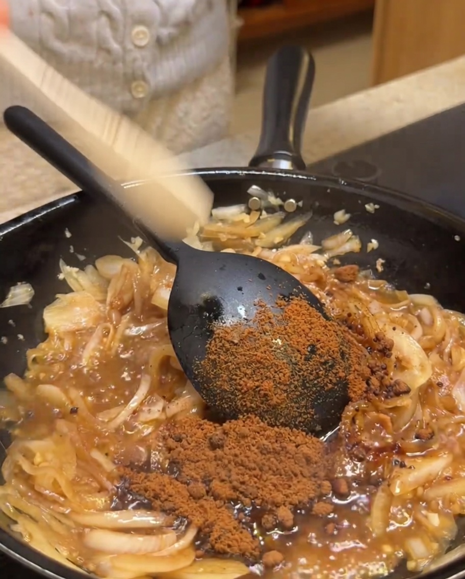 Brauner Zucker wird in eine Pfanne mit den sautierten Zwiebeln und Aromaten gestreut.