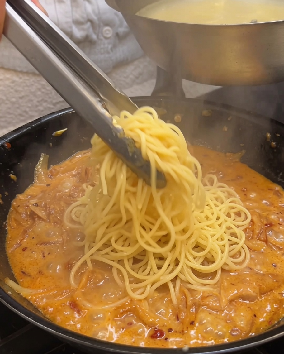 Gekochte Spaghetti werden mit einer Metallzange in einer cremigen Zwiebelsauce geschwenkt.