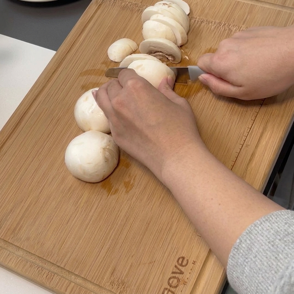 Eine Hand schneidet mit einem Kochmesser Champignons auf einem Bambusbrett in dünne Scheiben.
