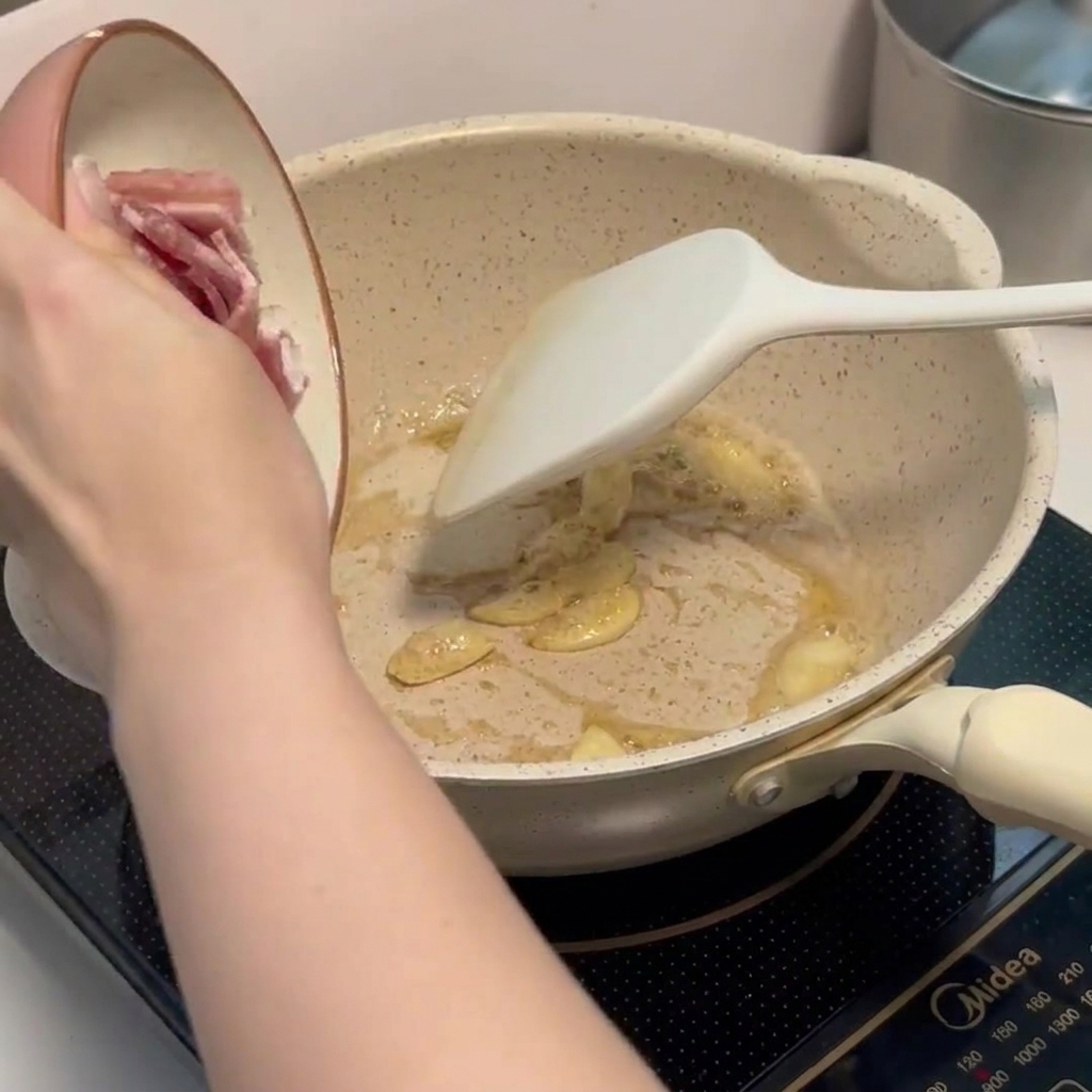 Kleine Speckstücke braten in einer Pfanne zusammen mit duftendem Knoblauch.